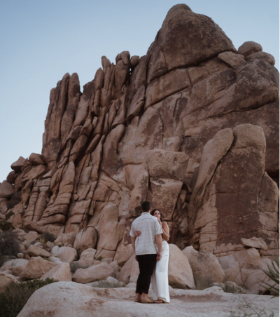couple is standing on a rock at sunrise looking out into the distance. they are dressed in a white jumper and the groom in a tan shirt and black pants.