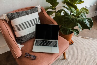 Mac book is sitting on a red sofa next to a white and grey pillow, green plant in the background