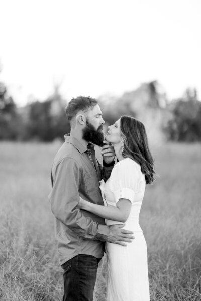 A black and white portrait of a couple in a grassy field, sharing an intimate moment as the woman gently lifts her partner's chin, capturing timeless romance and connection