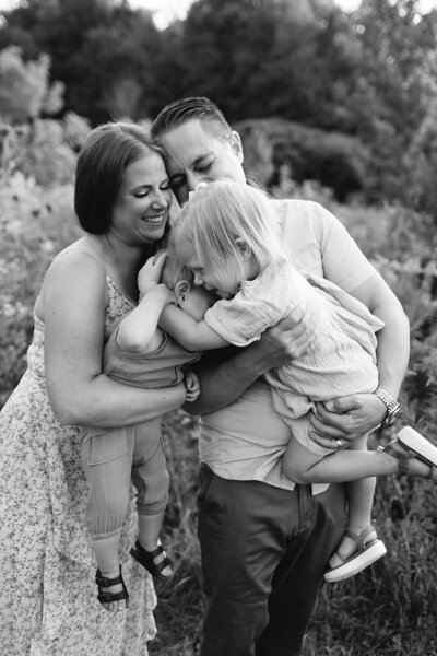 A woman sits on a deck, embracing two children. One smiles widely while hugging her, the other sits calmly on her lap. Captured like a scene from a family film by a talented Toronto family photographer, the background is dotted with trees and outdoor furniture.