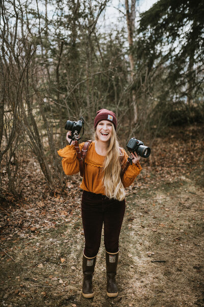 photo of Donna of Donna Marie Photography standing and smiling with her camera while wearing a black and red checkered flannel and black carhartt beanie