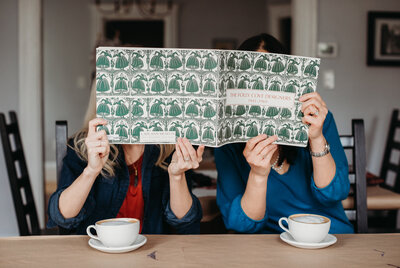 women hide behind magazine with lattes