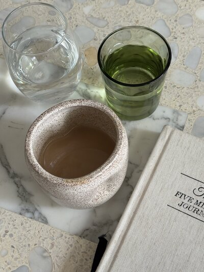 Ceramic mug with coffee, glasses of water and green juice on a marble table. Beige journal with "Five Minute Journal" text, creating a calm, organized setting.
