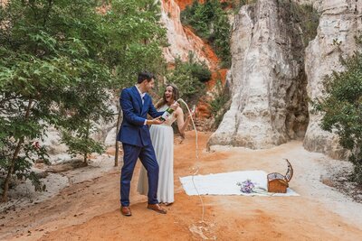 A bride and groom look at each other smiling and laughing happily after popping a bottle of champagne after their elopement. They are standing at the bottom of a canyon surrounded by red dirt, green trees and white and orange canyon walls. The bride is in a flowy white dress and the groom is in a navy blue suit.