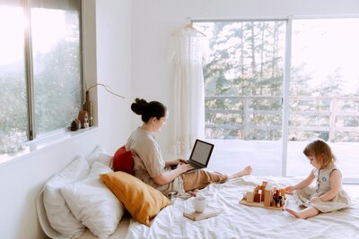 Mom sitting in bed on her laptop with her daughter playing next to her. 