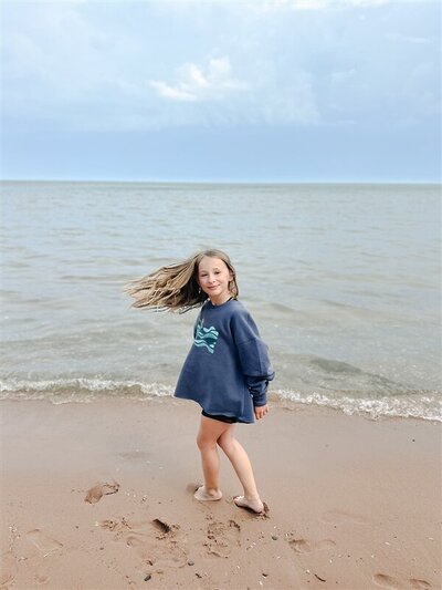 Young girl with brown hair wearing a large blue sweatshirt spins toward the camera on shore of Lake Superior