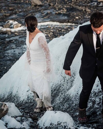 A bride and groom trek on the glacier in their formal wear during their two day luxury Alaska Glacier Elopement.  The bride has her dress slightly lifted as they walk up the hill, she is turned and looking way and you can see a peek of white, fur lined boots with ice cleats on them as they walk.  