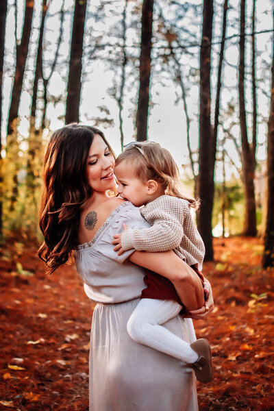A woman is standing holding a toddler in her arms and looking down smiling at the toddler. They're in a forested area.