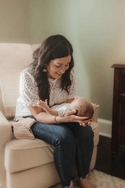 Mother smiling down while cradling her newborn in a nursery chair in Macon, GA.