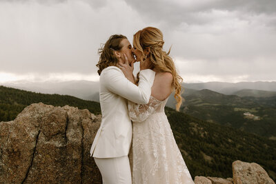 photo of brides kissing while standing on rock cliff with mountains in the background taken by Adventure elopement photographer in boulder
