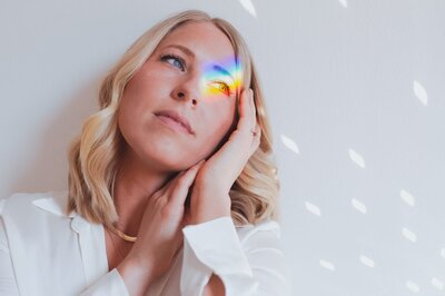 Woman with short, blonde, wavy hair softly rests her head on prayer hands with rainbow and discoball reflections on her face