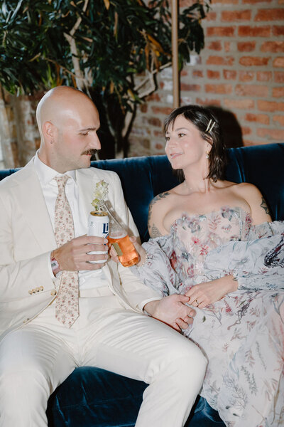 Bride in earthy, flowy, watercolor-printed dress sits beside groom on wedding day while clinking together their drinks at reception.