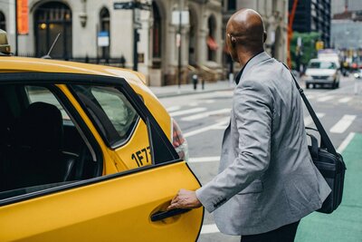 A man in a light gray suit opening the door of a yellow taxi on a city street.