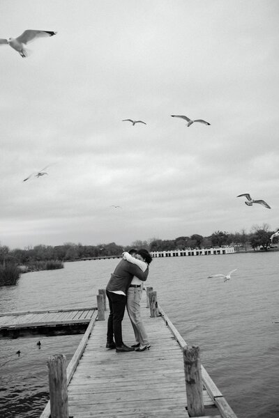 A romantic black and white engagement photo of a couple embracing on a dock with birds flying overhead. A cinematic and timeless love story captured by the water.