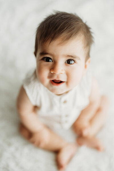 six month old baby boy lays on his belly on a white rug looking out the window while wearing a white diaper cover for his six month old Denver Mini milestone session