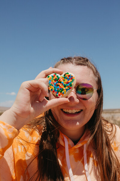 Girl in orange hoodie smiling while holding a cookie to her eye.