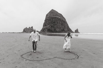 Black and white images of a couple drawing a heart in the sand in front of Haystack Rock at their Canon Beach Oregon Elopement.