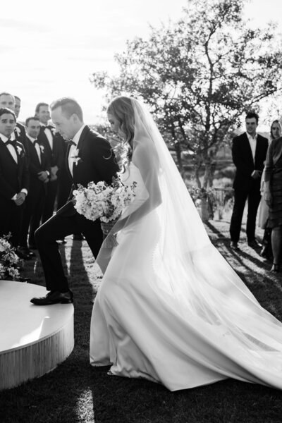 A groom assists his bride stepping up onto the platform for their ceremony in black and white. She has a long, trailing gown and veil that glows with the light passing through.