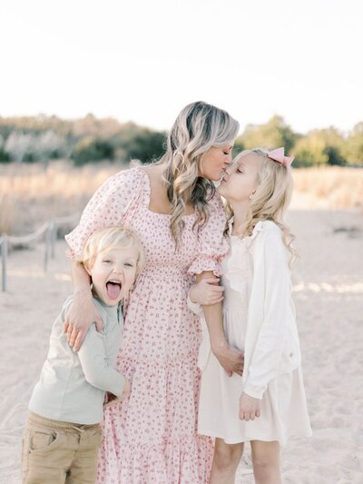 Blonde woman wearing pink floral dress kisses younger daughter and holds son who is making a  silly  face.