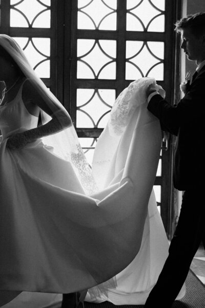 black and white overlay of bride being assisted by someone in a suit, white wedding dress train being carried during wedding photography session