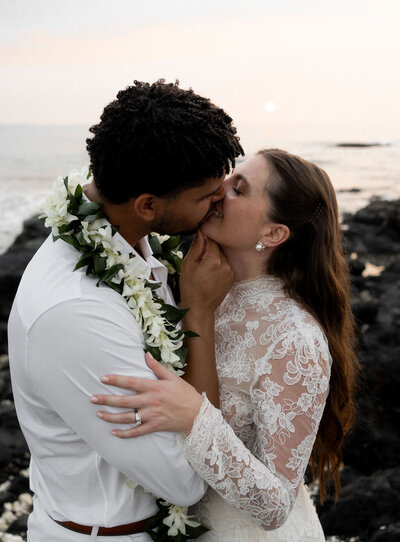 Bride and groom sharing a romantic kiss, with the bride elegantly dressed in a long-sleeve lace wedding gown and the groom wearing a Maile lei with a white orchid twist, capturing a timeless moment from their luxury destination wedding