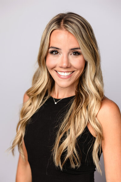 Corporate headshot of a smiling woman with long blonde hair wearing a black top on a light background, Long Island professional headshots.
