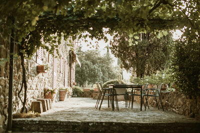 Outdoor patio with a stone floor, featuring a metal table and chairs under a lush vine-covered pergola. Potted plants and trees surround the area, creating a serene and rustic atmosphere—discover more tranquil spaces at https://epicsandstravel.com/about.