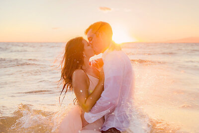 Couple kiss in the ocean as a wave splashes on them during their proposal portraits in Maui by Love + Water