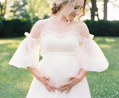 A blonde pregnant mother holds her belly while wearing a white flowing dress during her maternity session in Maryland