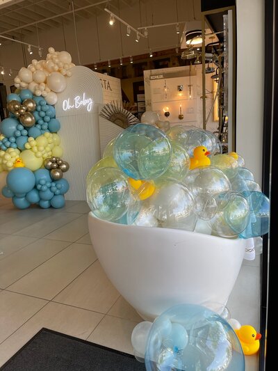 Balloon installation inside of a bathtub with rubber duck theme. Backdrop in the background with a second balloon installation in the colors blue, pale yellow, and gold. 