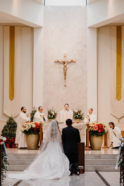 Bride and groom kneel at the alter in beautiful bright Catholic Church. Photo taken by  Orlando Wedding Photographer Four Loves.