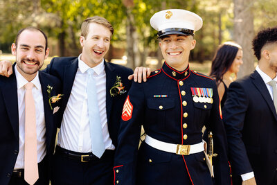 A group of people outdoors, smiling and walking together, is beautifully captured by a Brainerd Lakes Area Photographer. One person is in a U.S. Marine dress uniform with medals, while the others wear suits. Trees and greenery provide a lush backdrop to this memorable scene.
