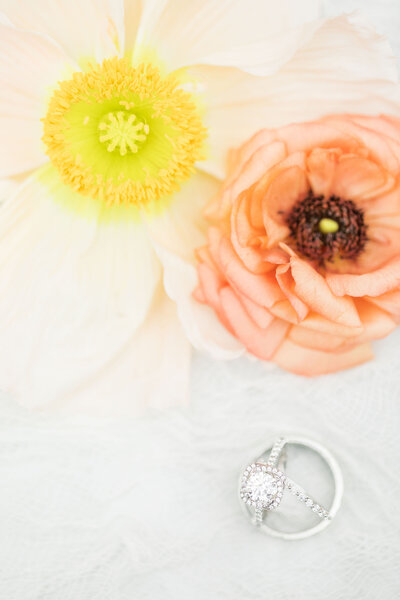 Macro shot of the wedding bands and engagement ring laid upright on a styling mat with a light pink peony and a coral peony at the Ivory Oak wedding venue in Texas