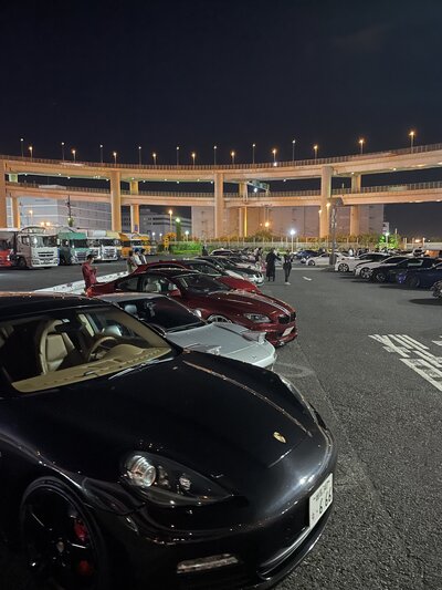 A row of luxury supercars including a Maserati and Ferrari parked under a vibrant Tokyo sky at the iconic daikoku Parking Area meet-up spot.
