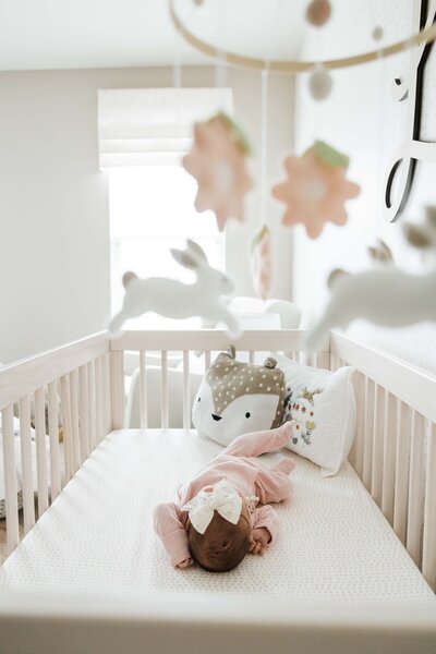 A baby rests in a crib with a mobile and plush toys nearby, a perfect scene for in-home newborn photography.