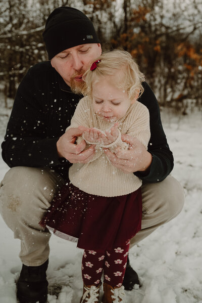 Dad holding daughters hands while they are both blowing snow from their hands in Maryland
