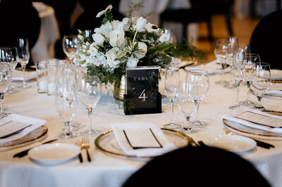 Elegant wedding tablescape at the Palm Event Center in Pleasanton, CA, during a spring reception. The design features a white flower centerpiece, black and gold table numbers, and luxurious gold place settings.