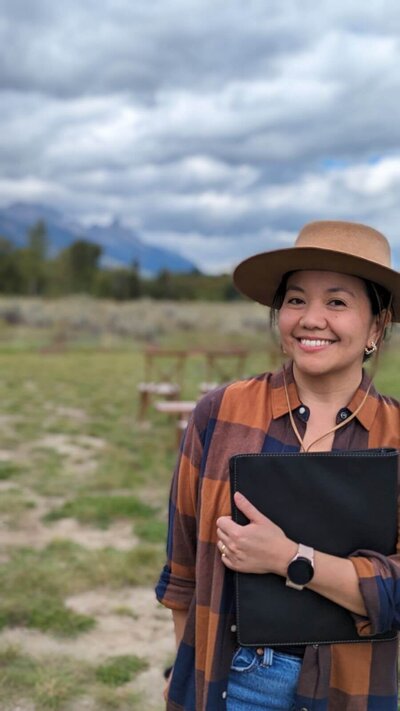 Idaho, Wedding Planner, Maria Tzompa with Felicity Events smiles at the camera outdoors with mountains and cloudy skies in the background, representing her vendor profile for the Crush Collective wedding show.