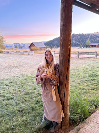 Marcie Muensterman is in a large robe holding a mug and leaning against a tall wooden post. The background shows a serene Montana sky and barn.