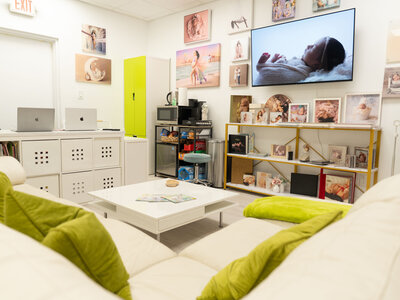 Cozy modern room with white shelves, laptops, green cushions, and baby photo art. A large TV shows a sleeping baby. Warm, inviting atmosphere.