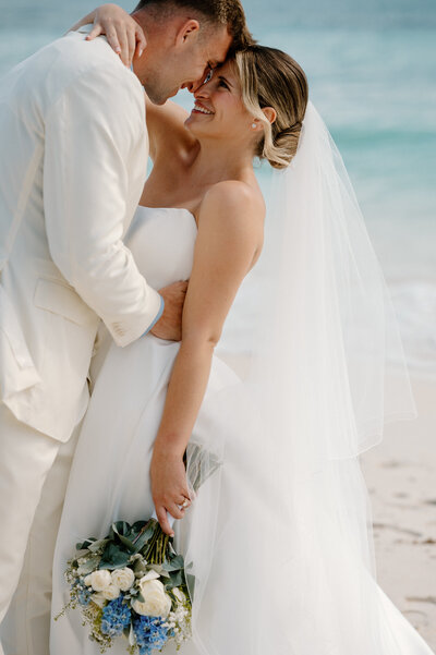 Bride and groom embrace on the beach in Mexico, smiling against turquoise water during their destination wedding.