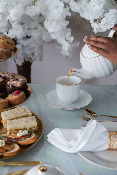A person's hand pouring tea from a white and gold teapot into a teacup. A tiered tray of sandwiches and desserts is visible in the background.
