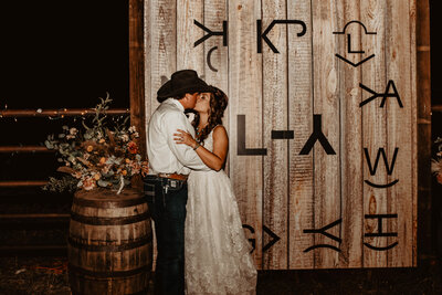 bride and groom kissing in front of branding wall at their ranch wedding
