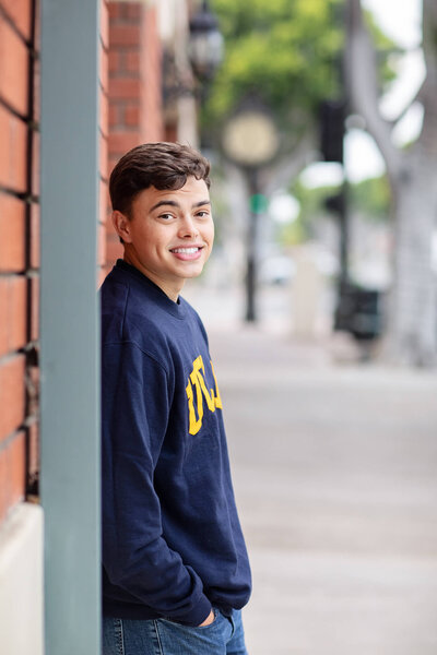 Young man in a black shirt and jeans sitting on the ground with arms crossed in front of a brick wall