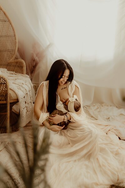 A woman in a cream dress sits on the floor in a light-filled room, breastfeeding a baby wrapped in a beige cloth. The room, perfect for newborn photography, is decorated with soft fabrics and a wicker chair, capturing an intimate moment that would be cherished by any Macon, GA family.