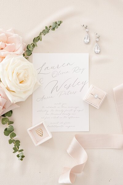 ivory and blush pink wedding invitation suite styled with florals, ribbon, a ring box, earrings, and a pastel pink backdrop.