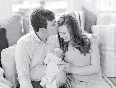 A beautiful black and white image of new parents embracing their newborn baby on a cozy couch. The father gently kisses the mother’s forehead as she smiles, holding their little one with love.