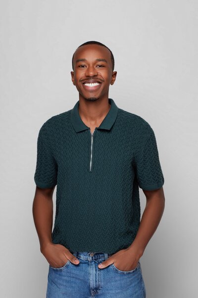 Portrait photo of Dennis, a young black man with short hair. He has his hands in his jean pockets and is smiling. He is wearing a dark teal polo shirt