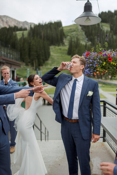 A groom drinking a shot at the base of Arapahoe Basin with his wife behind him