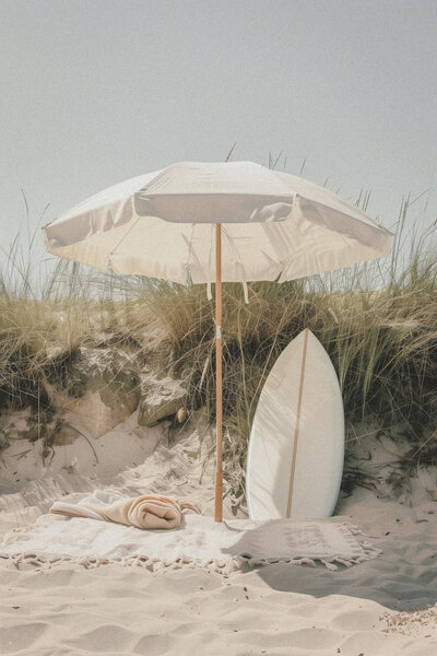 White beach umbrella with blanket white boogie board towel and sitting in front of grassy dune at beach